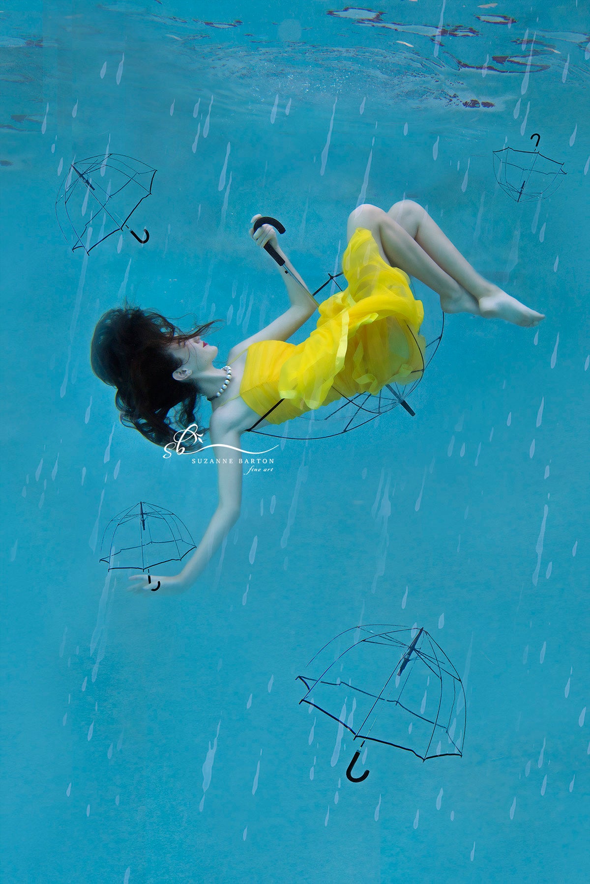 Dark-haired woman wearing a bright yellow dress floating underwater while sitting in a clear umbrella. Raindrops fall through the aqua blue water.