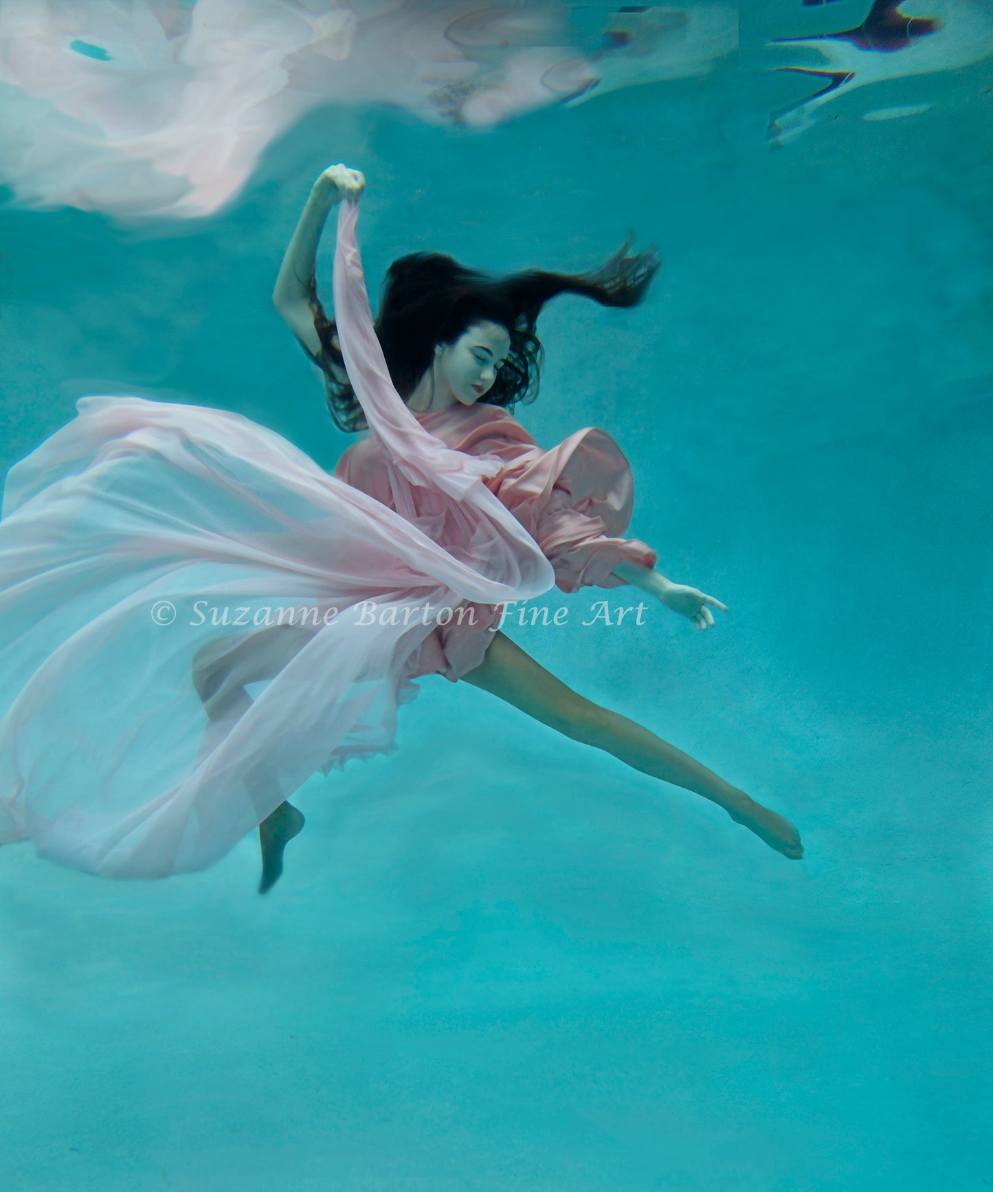 A striking woman with long black hair in flowing pink dress underwater and surrounded by aqua blue.