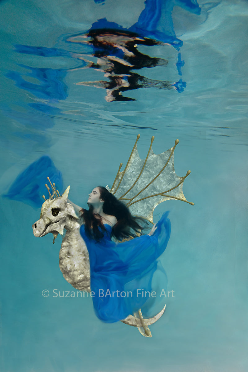 Fantastical image of a woman in a vibrant blue dress riding a gold magical seahorse dragon underwater.
