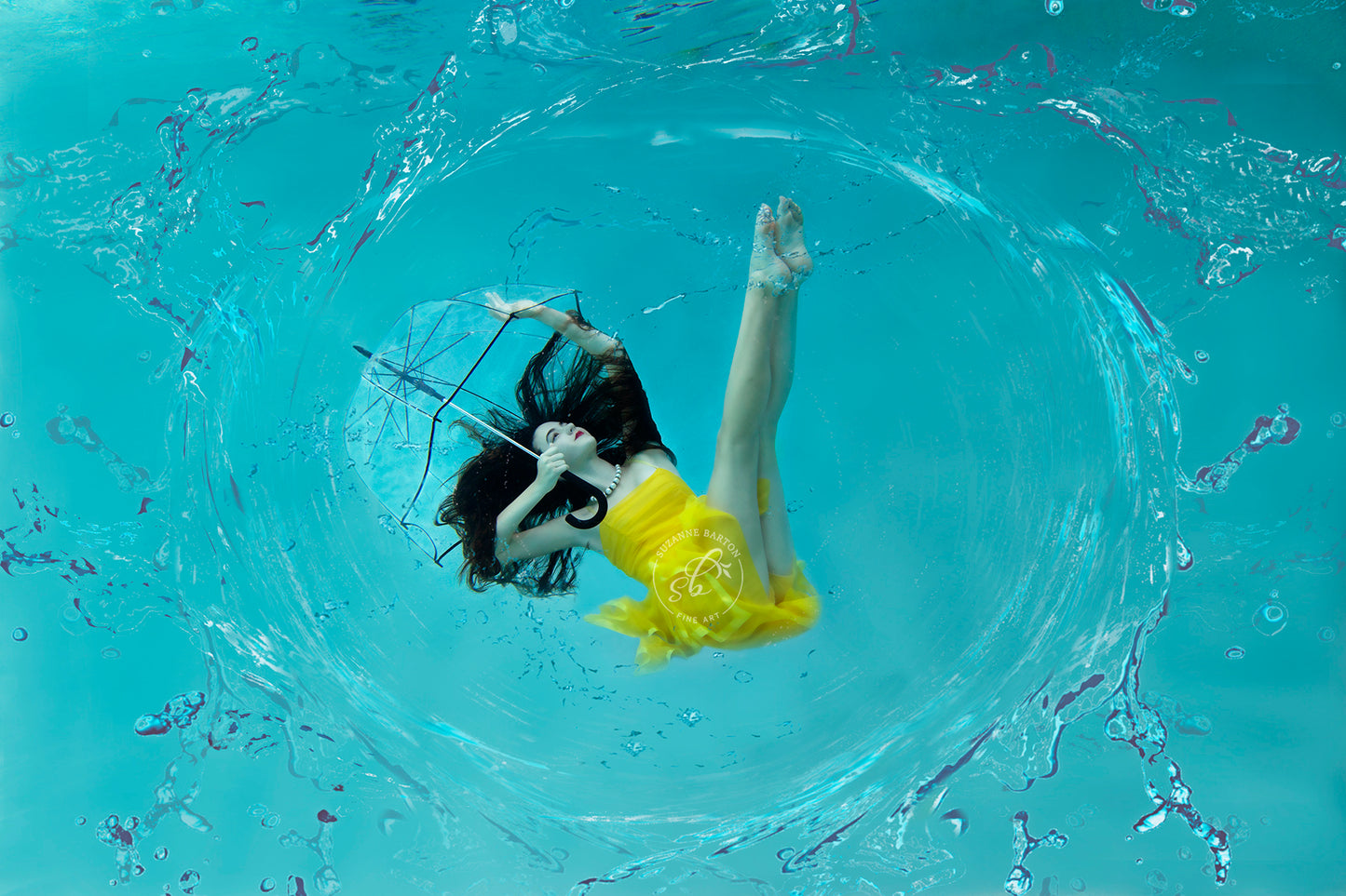 Fun underwater image of a beautiful woman with dark hair wearing a yellow dress floating while holding a transparent umbrella and surrounded by a water splash.