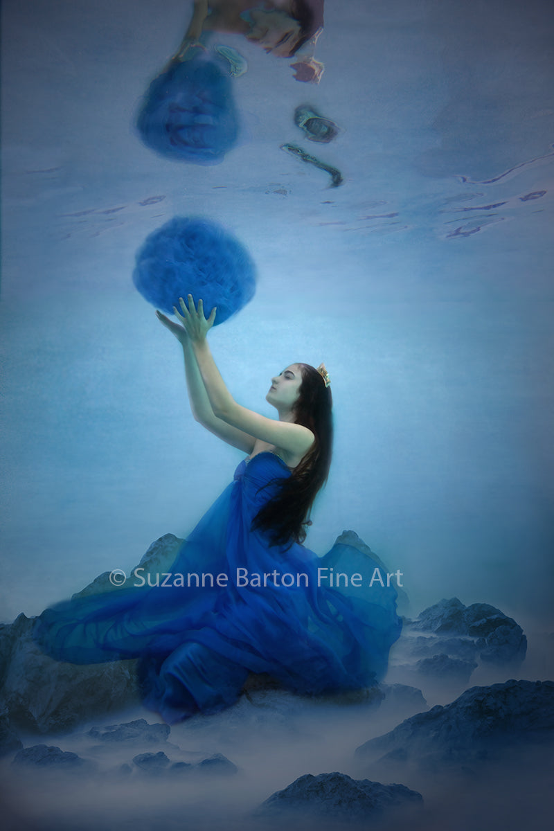 Woman underwater in vibrant blue dress flowing around her reaching up and holding a blue soft sphere.