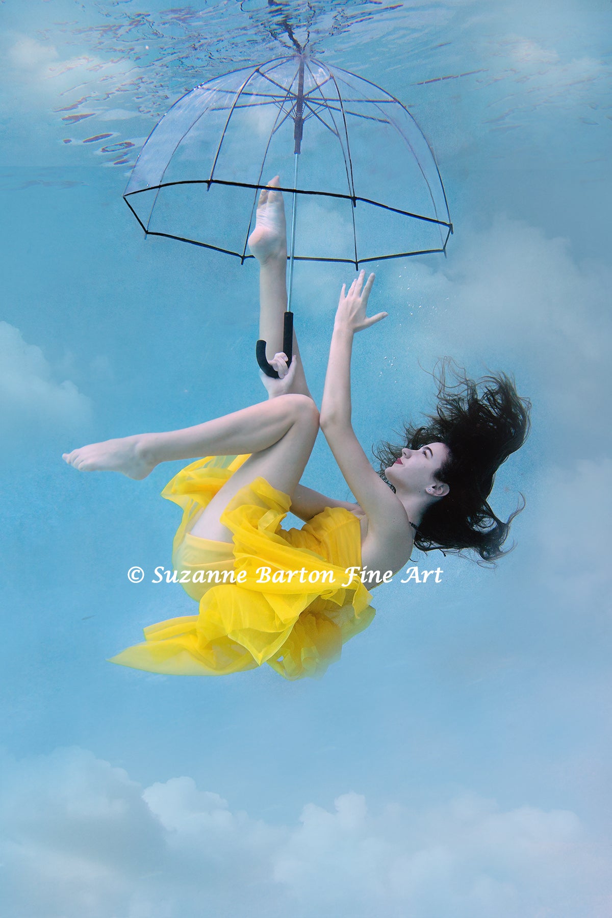 Dark-haired woman in a bright yellow dress holding a clear umbrella floats in a dancer pose while surrounded by aqua blue water.