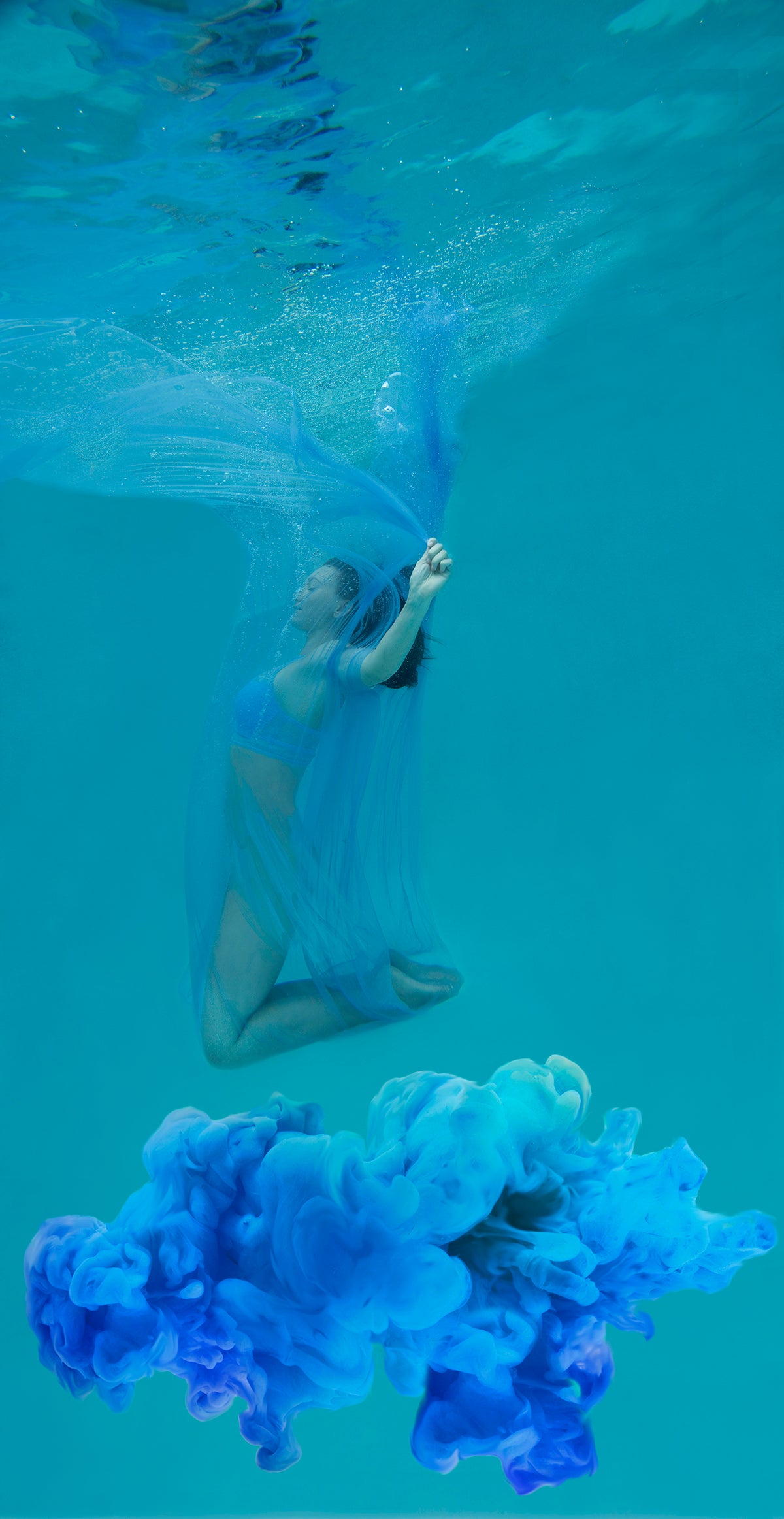 Woman floating underwater covered in a transparent flowing blue scarf. She is above a cloud of beautiful blue cloud.