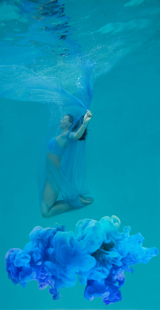 Woman floating underwater covered in a transparent flowing blue scarf. She is above a cloud of beautiful blue cloud.