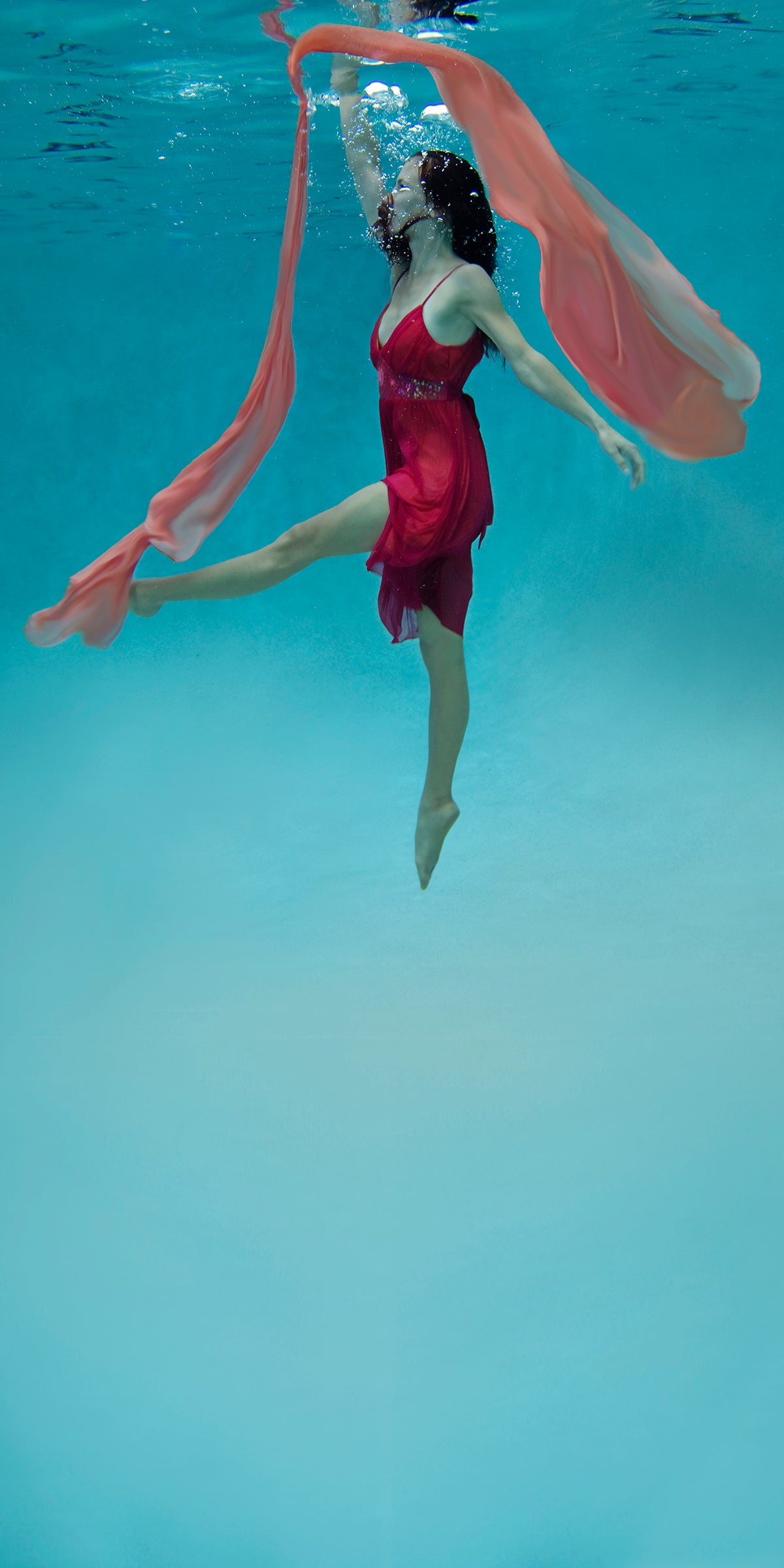 Woman wearing red dress floating underwater with a peach colored scarf overhead.