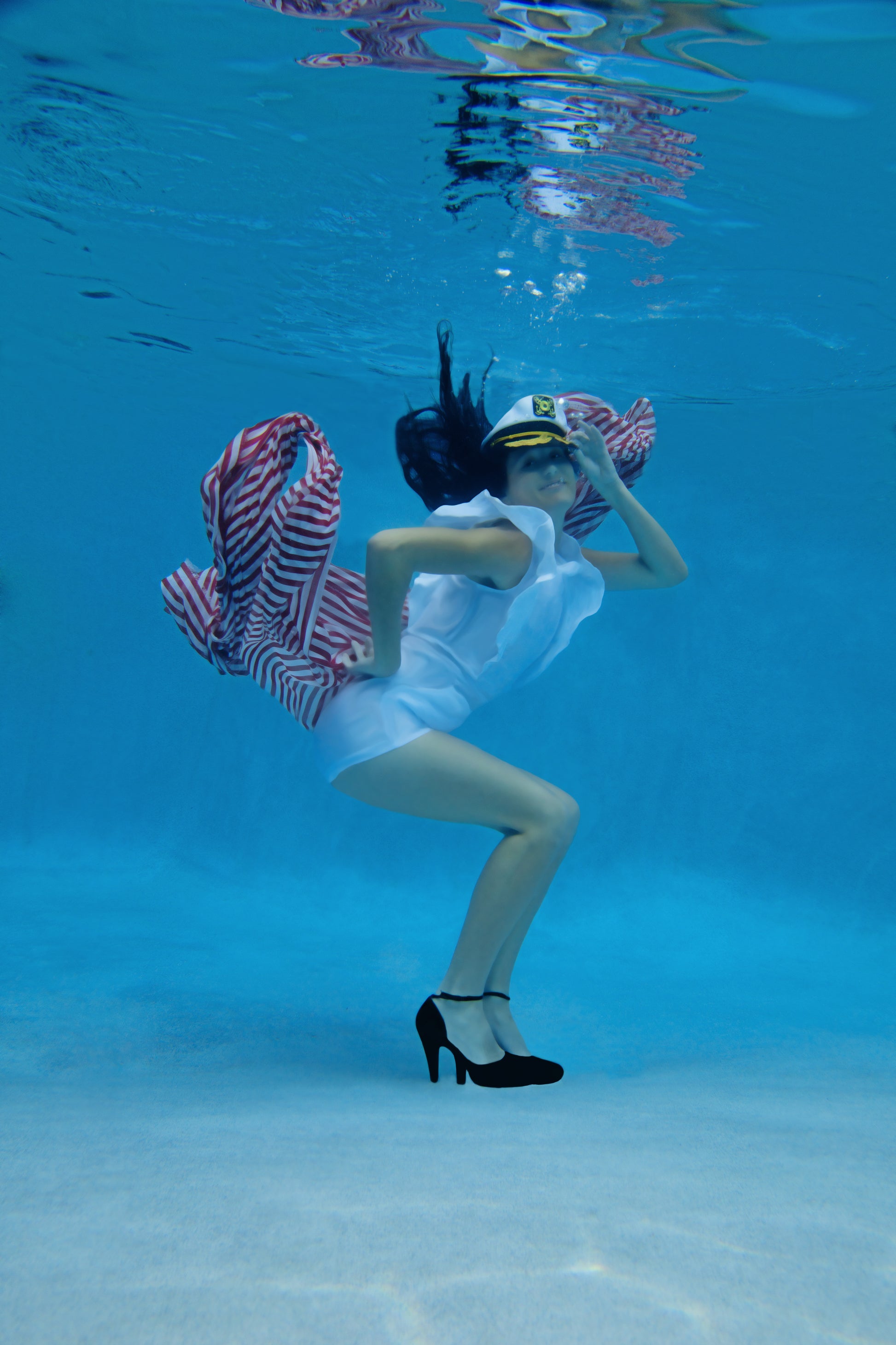 A stunning underwater fine art pinup-style image of a woman playfully wearing a sea captain's hat, white shorts, black heels and red and white striped scarf. 