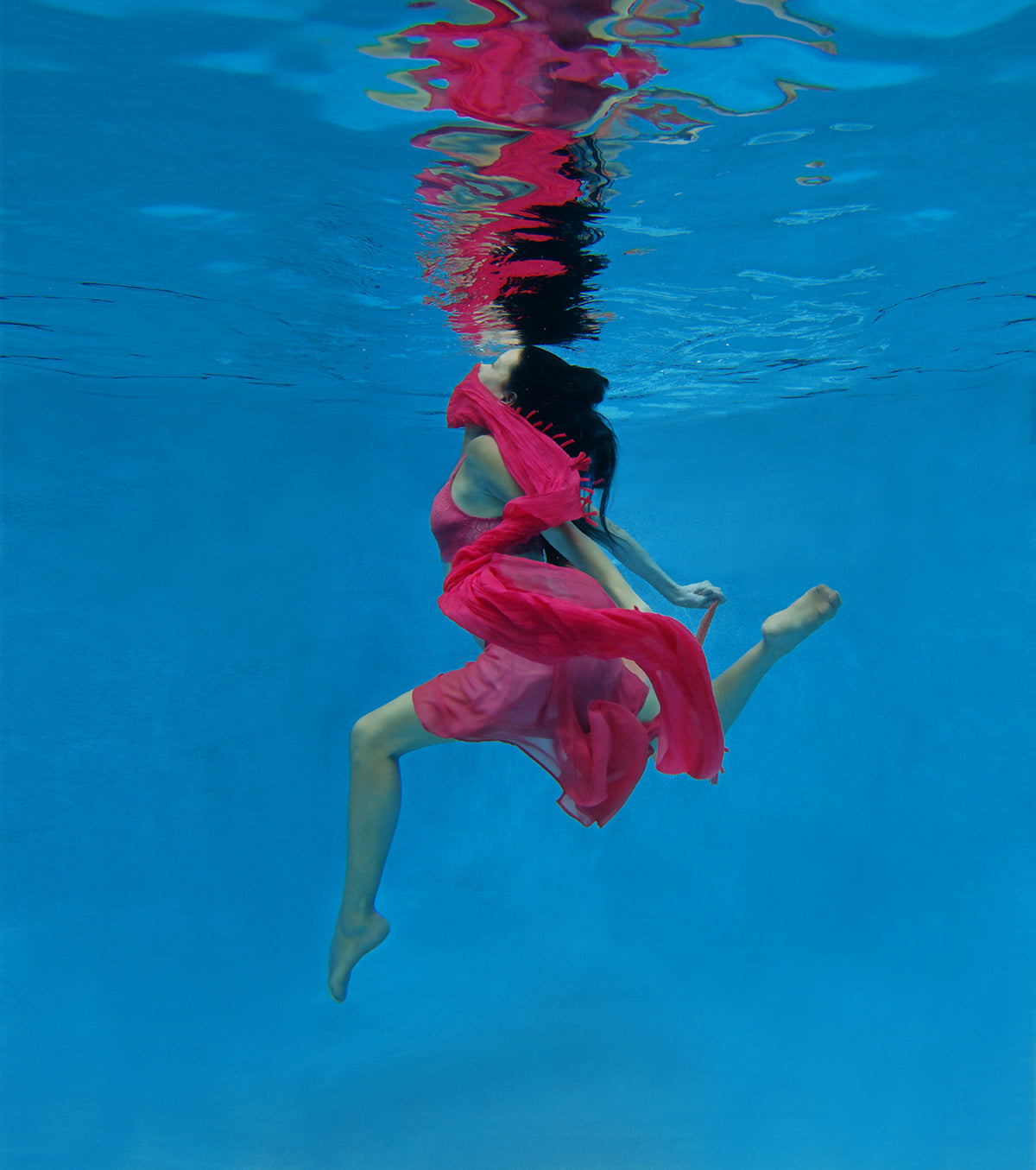 Arabian Nights shows an underwater scene of a young woman with a vibrant swirl of red fabric.The water is a vibrant aqua blue.