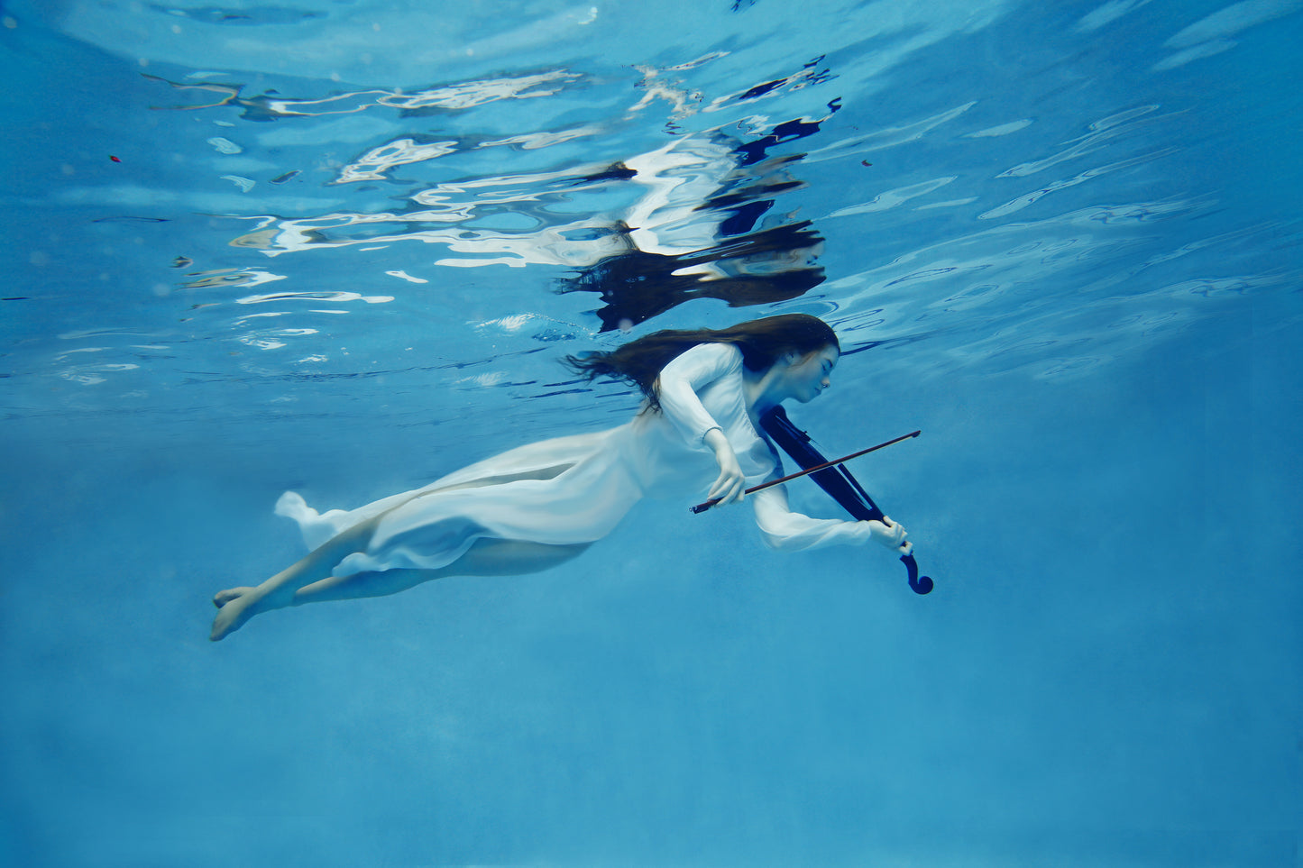 A woman in a flowing white dress playing the violin underwater portrays a stunning ethereal scene.